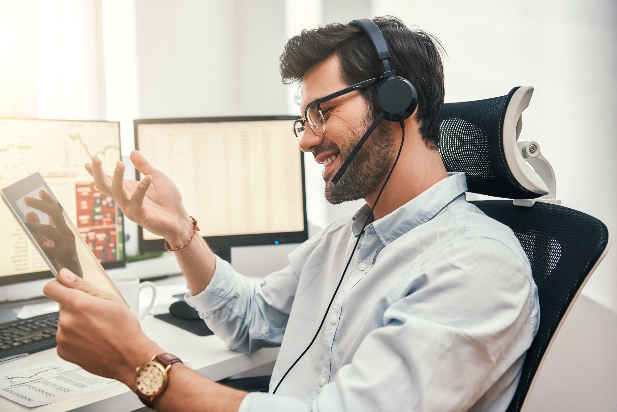 Happy trader. Successful bearded businessman in headset holding digital tablet, talking with client